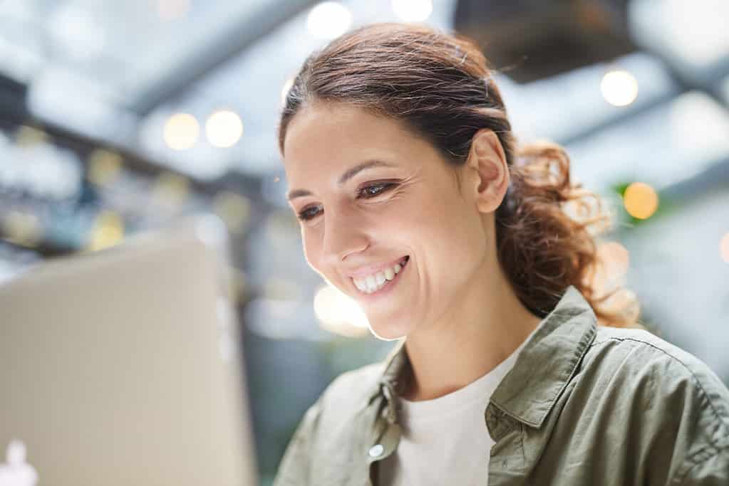 Close up portrait of smiling young woman using laptop in cafe sitting under glass roof on terrace